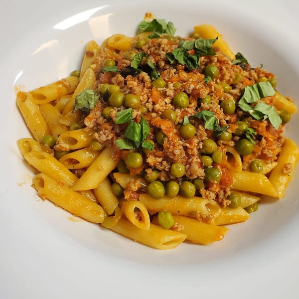 Penne pasta with a rich meat sauce, green peas, and fresh basil in a white bowl.